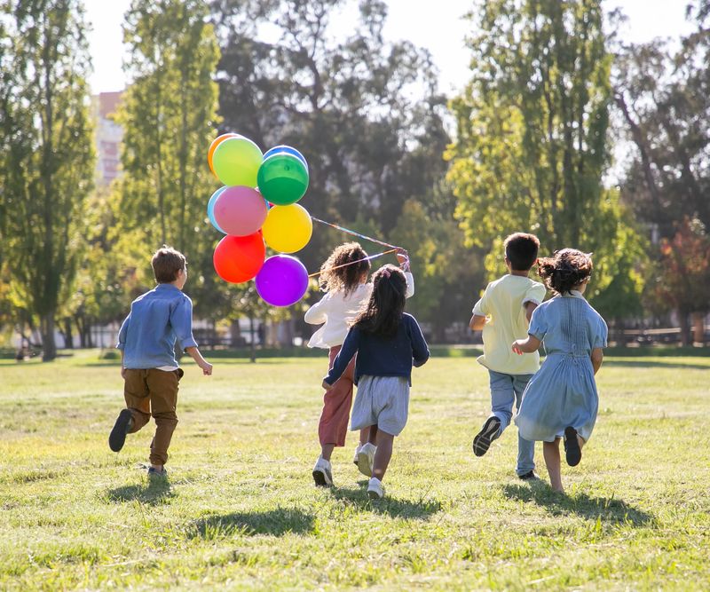 Children running across a park holding a bunch of balloons